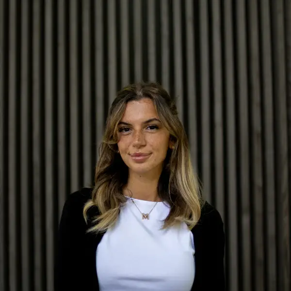 A woman with wavy, shoulder-length hair poses in front of a wooden vertical striped background, wearing a white shirt and black cardigan